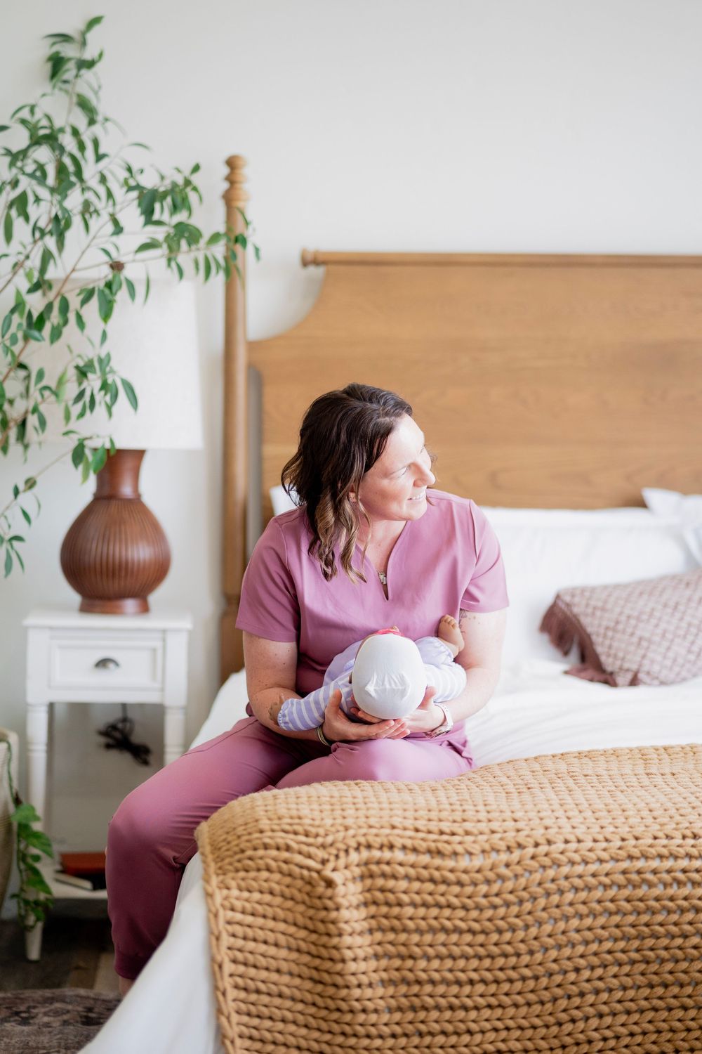 Female nurse in pink scrubs sitting on bed holding newborn, looking away in bright bedroom.