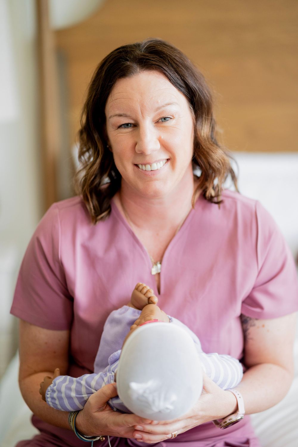 Smiling female nurse in pink scrubs holding newborn baby facing camera in warm bedroom.