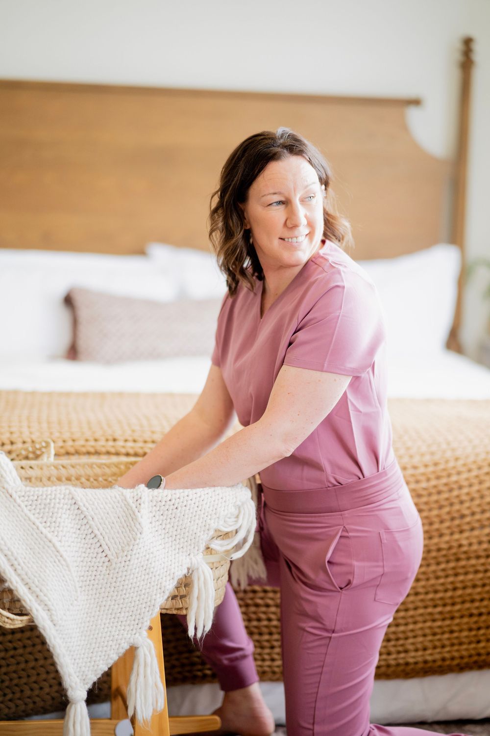 Smiling nurse in pink scrubs standing beside wooden bassinet with white knit blanket in bedroom.
