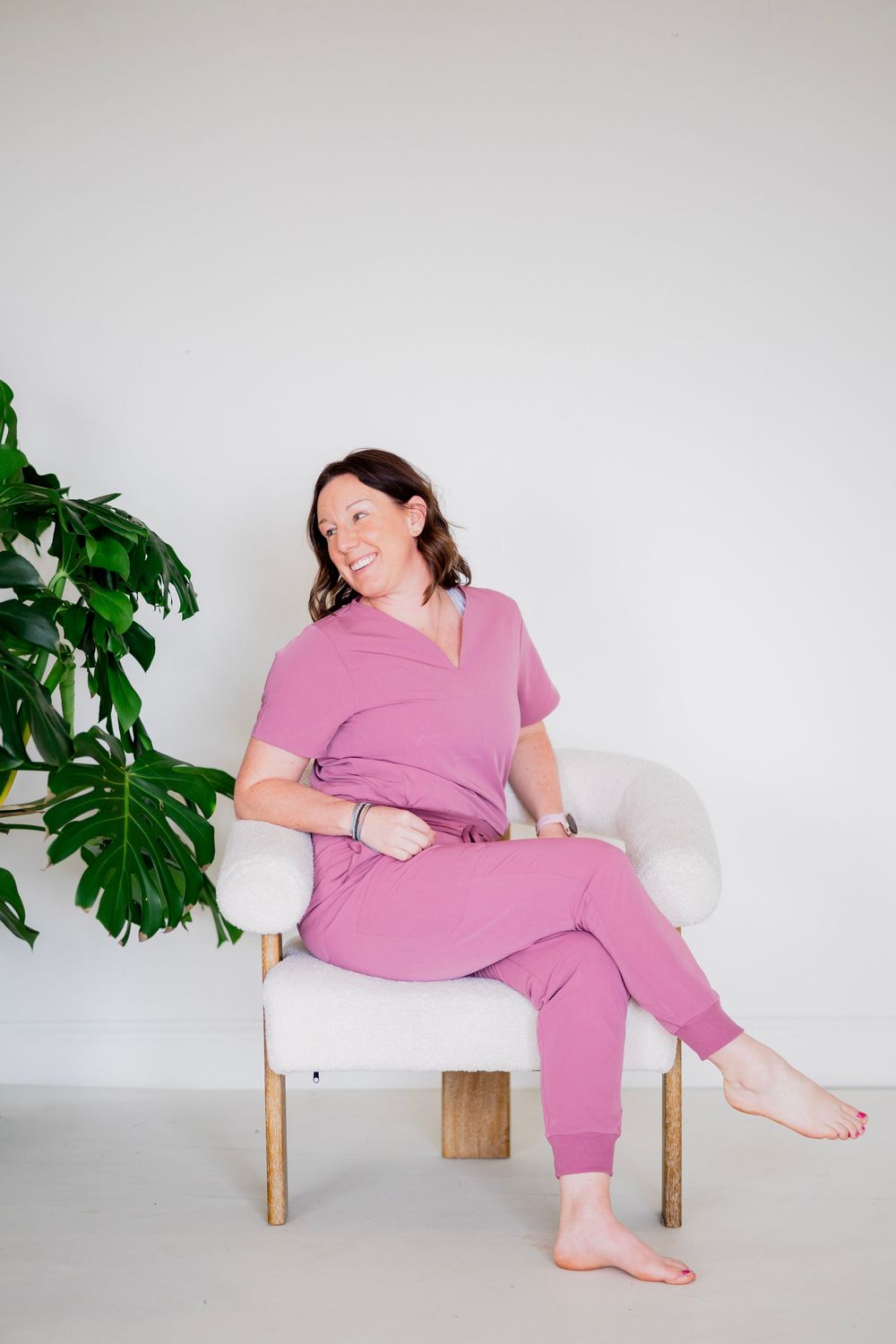 Woman in pink medical scrubs sitting barefoot in white chair beside monstera plant, laughing.