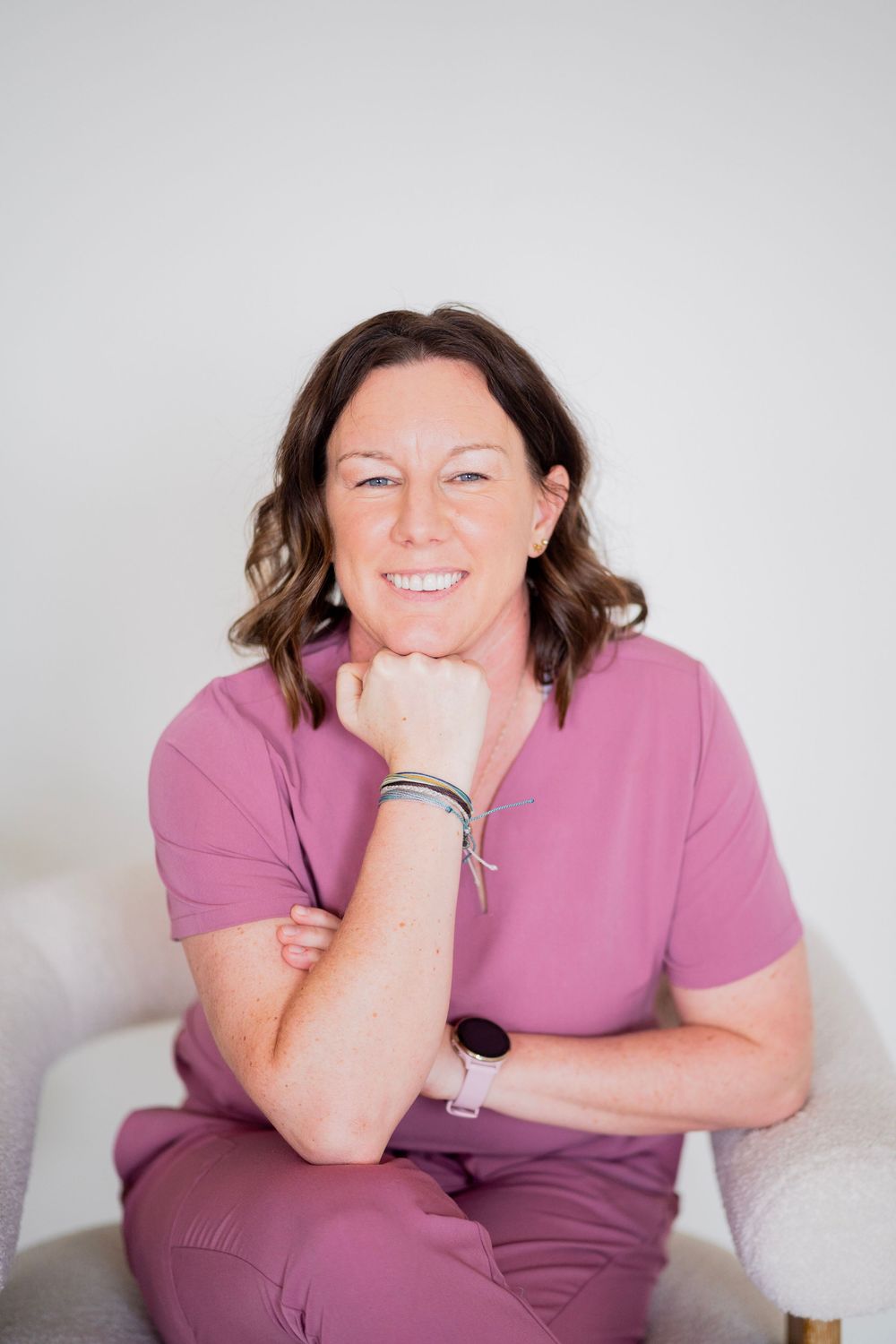 Smiling woman in pink scrubs sitting in white chair with chin resting on hand, portrait.