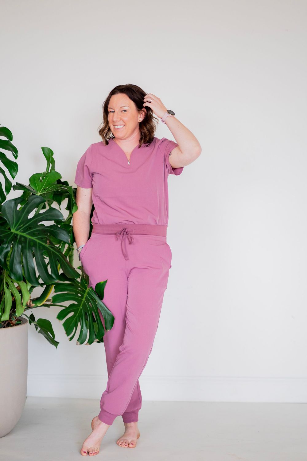 Woman in pink scrubs leaning against wall beside monstera plant, hand in hair, smiling.