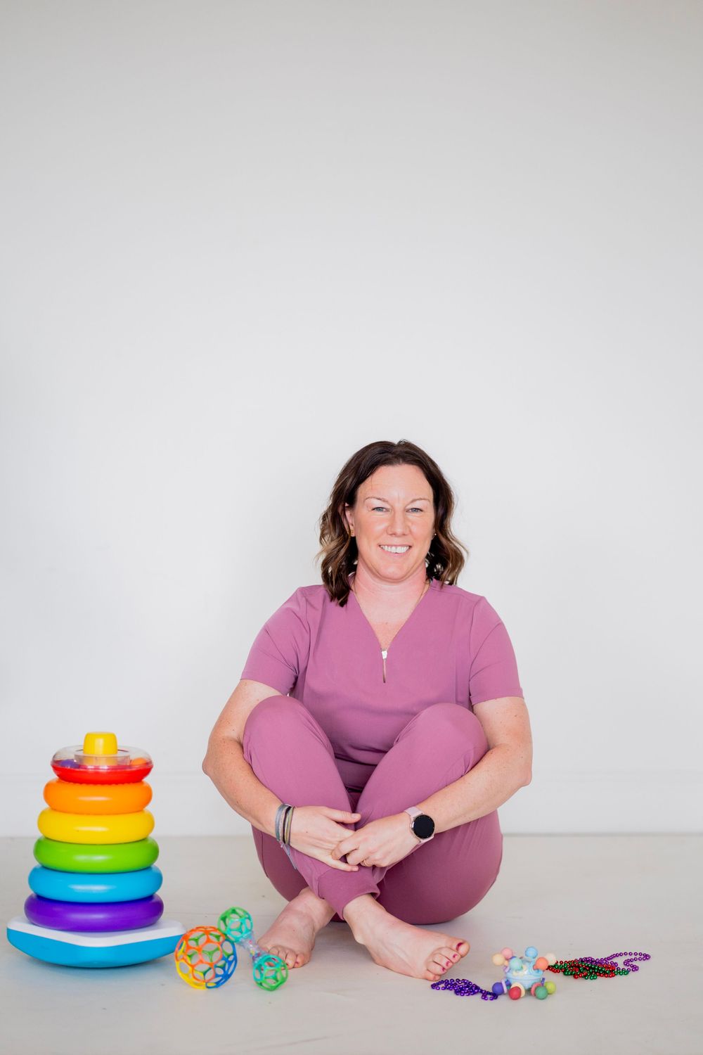 Woman in pink scrubs sits cross-legged on floor surrounded by colorful children's toys including a rainbow stacking ring toy.
