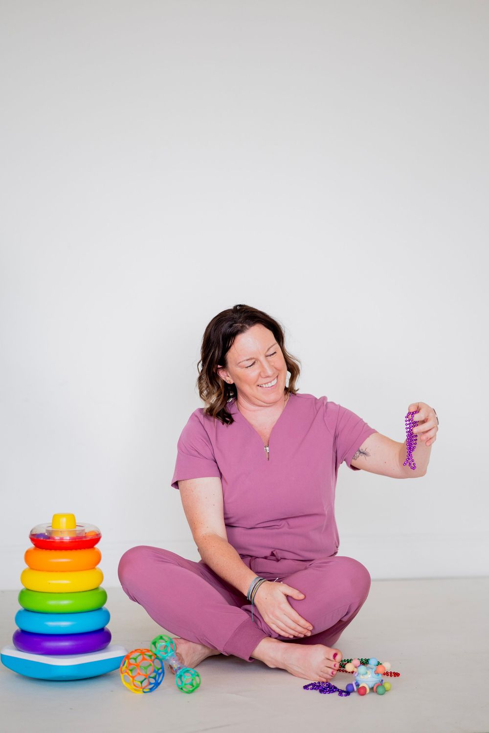 Woman in mauve scrubs sits cross-legged holding purple beaded necklace with colorful children's toys beside her.