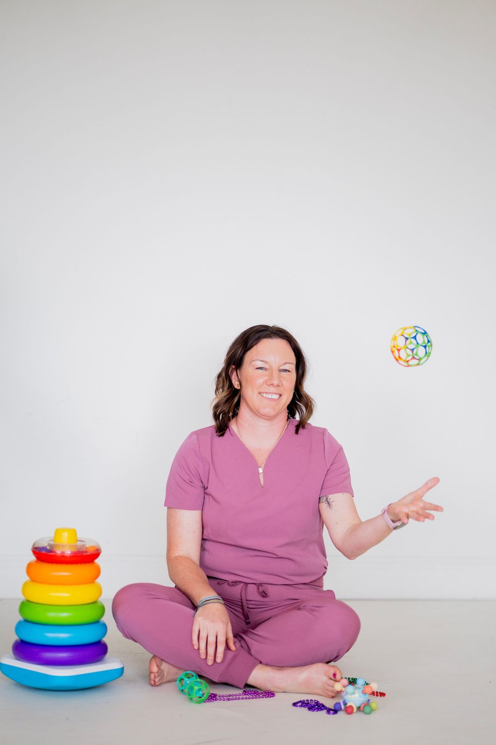 Smiling woman in pink scrubs tosses a colorful ball while sitting cross-legged next to a rainbow stacking toy.