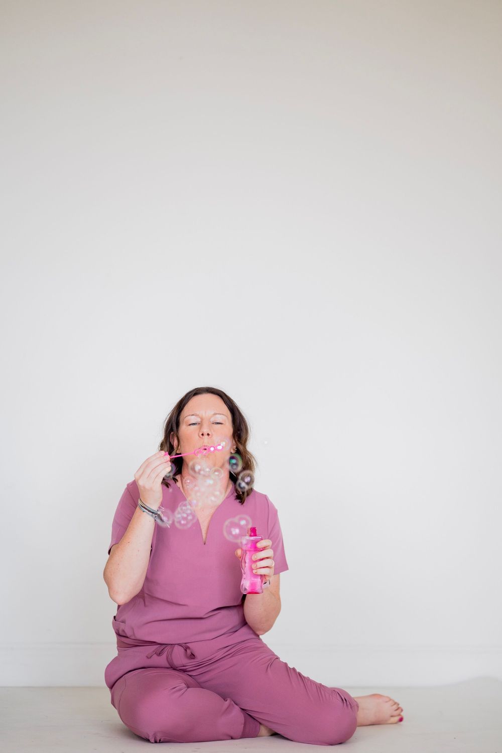 Woman in mauve scrubs sits on floor blowing bubbles from a pink bubble wand bottle against white background.