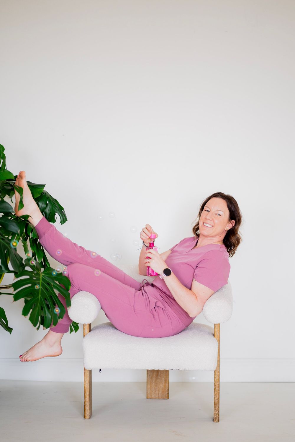 Laughing woman in pink scrubs reclines in white chair holding bubble wand next to large monstera plant.