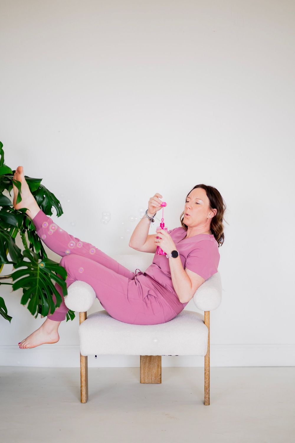 Woman in mauve scrubs reclines in white accent chair examining bubble wand next to a large monstera plant.