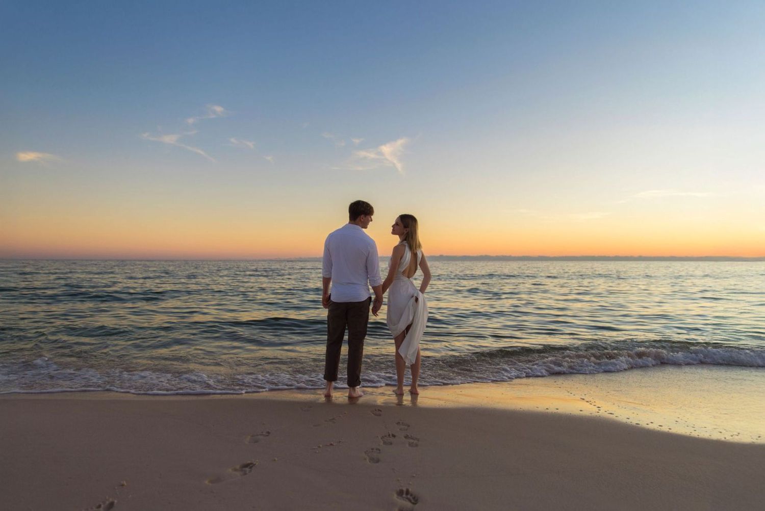 Couple holding hands on a sandy beach at sunset, with ocean waves and colorful sky in the background.