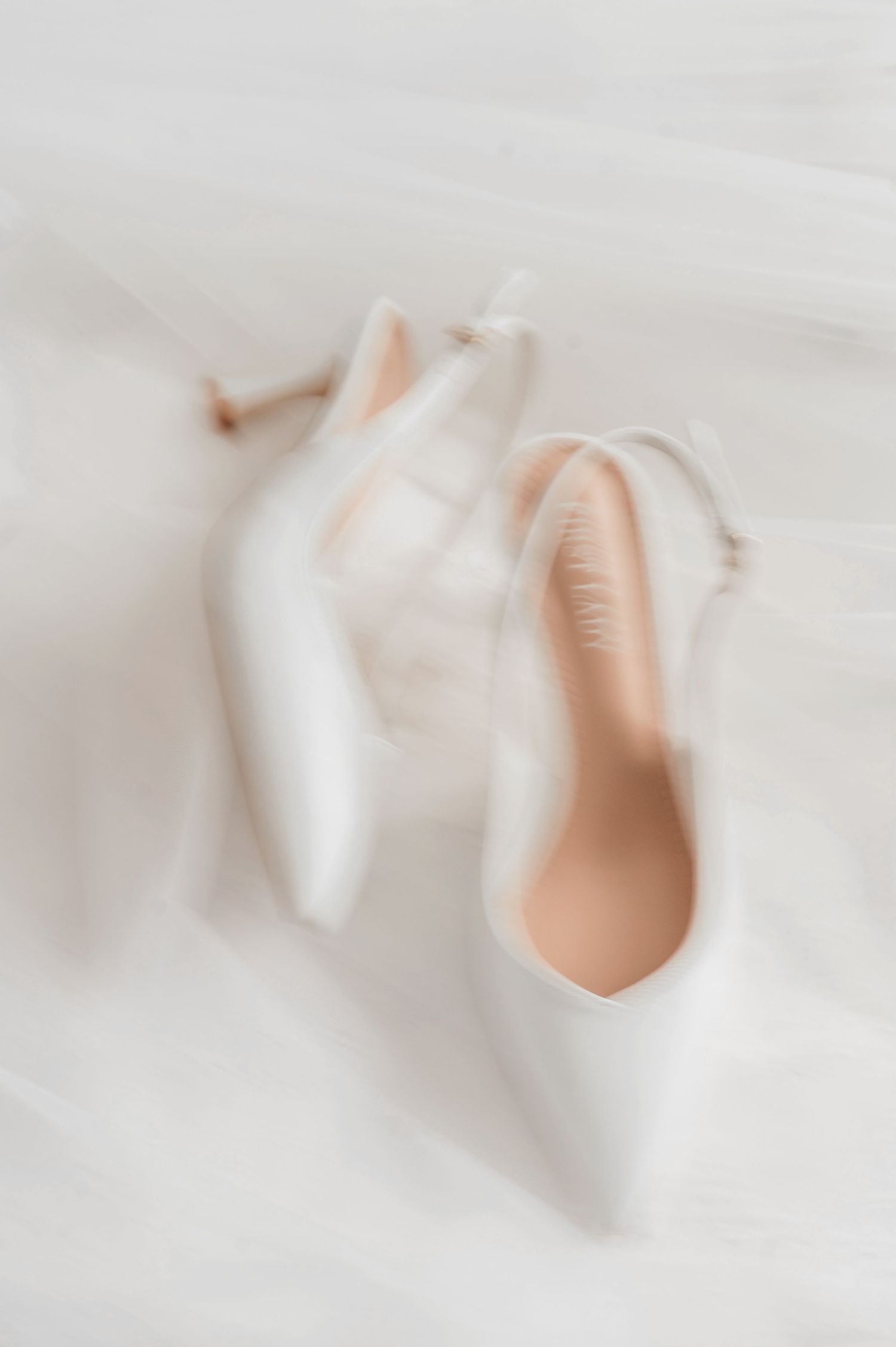 Soft focus artistic image of elegant white satin bridal heels with delicate ankle straps on veil.