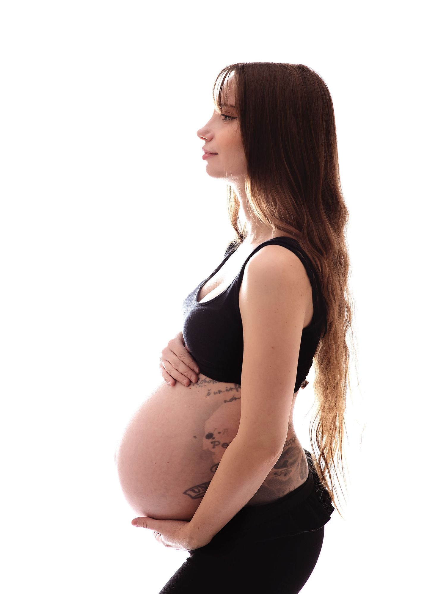 Pregnant woman in profile view cradling her bare baby bump, wearing a black crop top and leggings, with long brown hair.