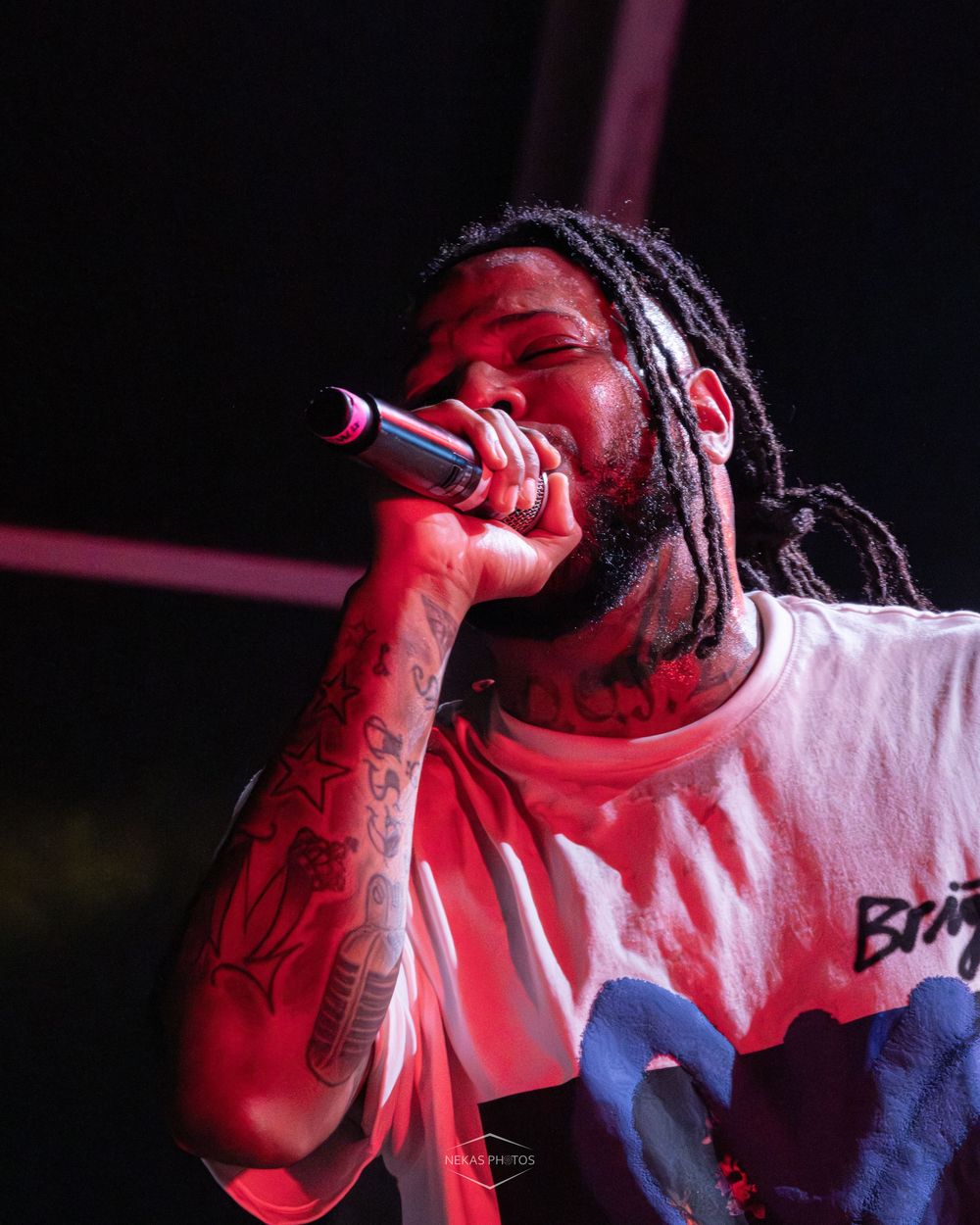 Tattooed rapper with dreadlocks performs on stage, holding a microphone under dramatic red lighting.