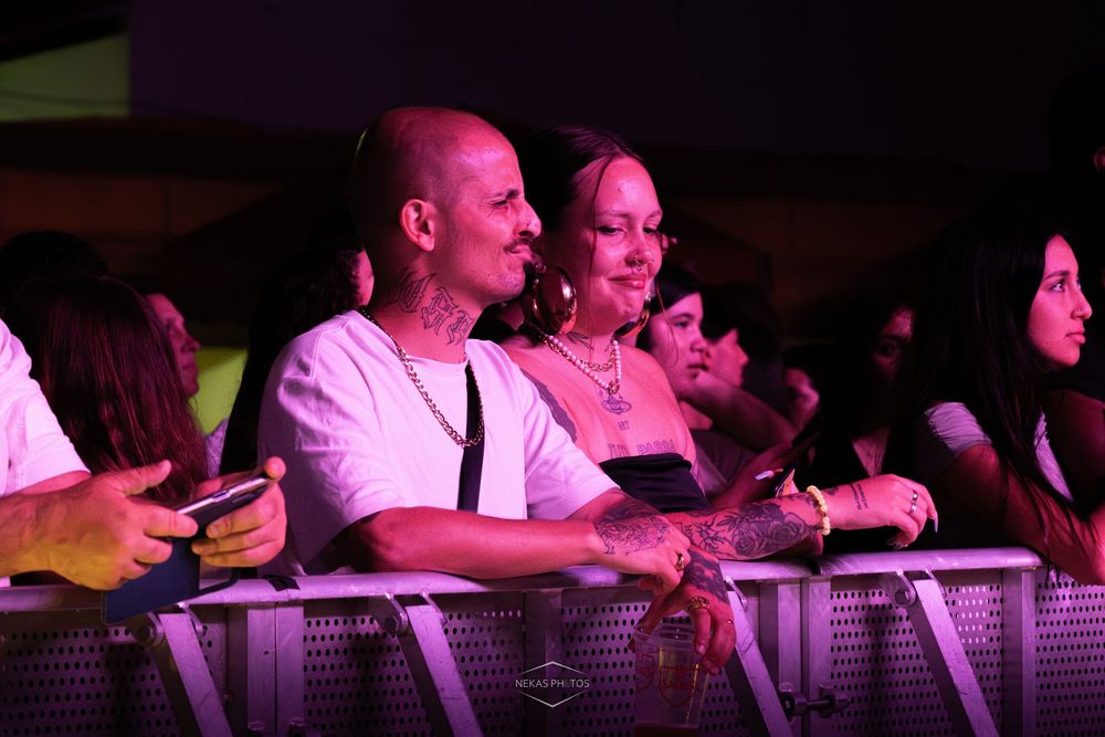 Tattooed couple smiling and leaning on a barrier at an outdoor concert under pink stage lights.
