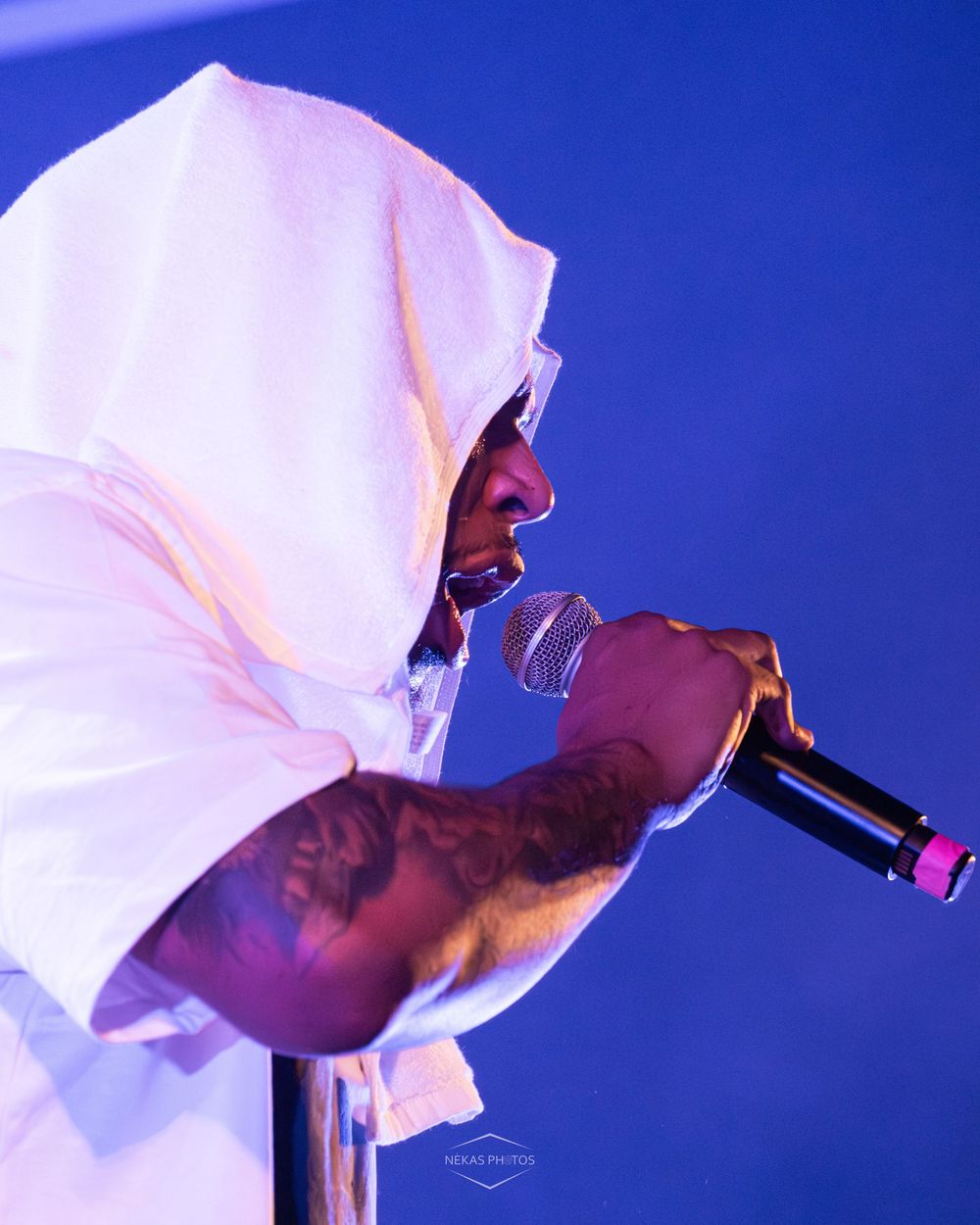 Tattooed male performer in white hoodie singing into microphone on stage with blue background.
