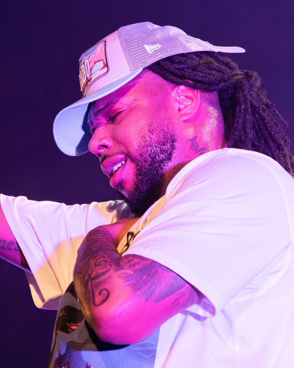 A tattooed man with dreadlocks and a white trucker hat performs on stage under pink and purple lights.