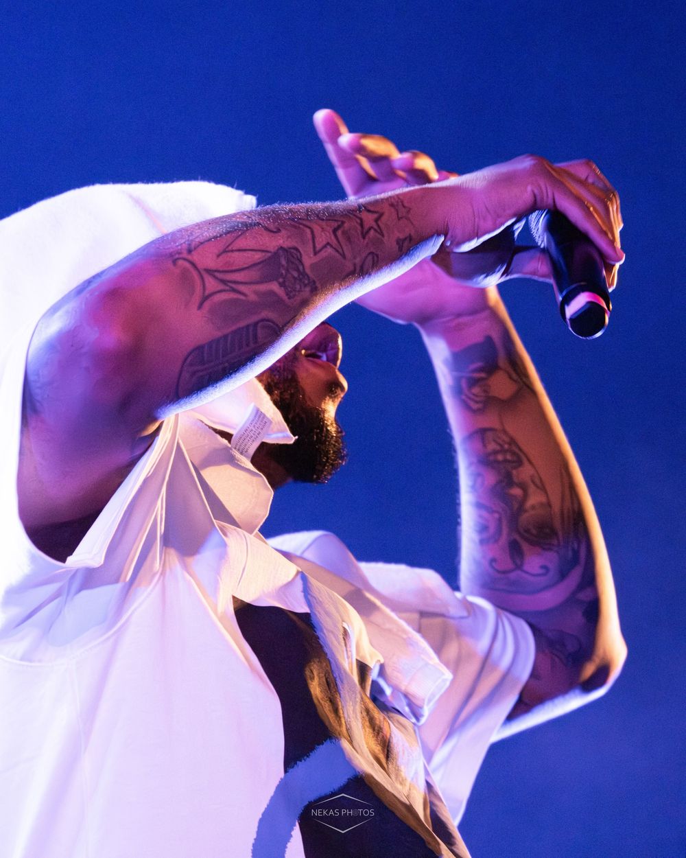 Tattooed male performer singing into microphone on stage, wearing white shirt and cap under blue stage lighting.