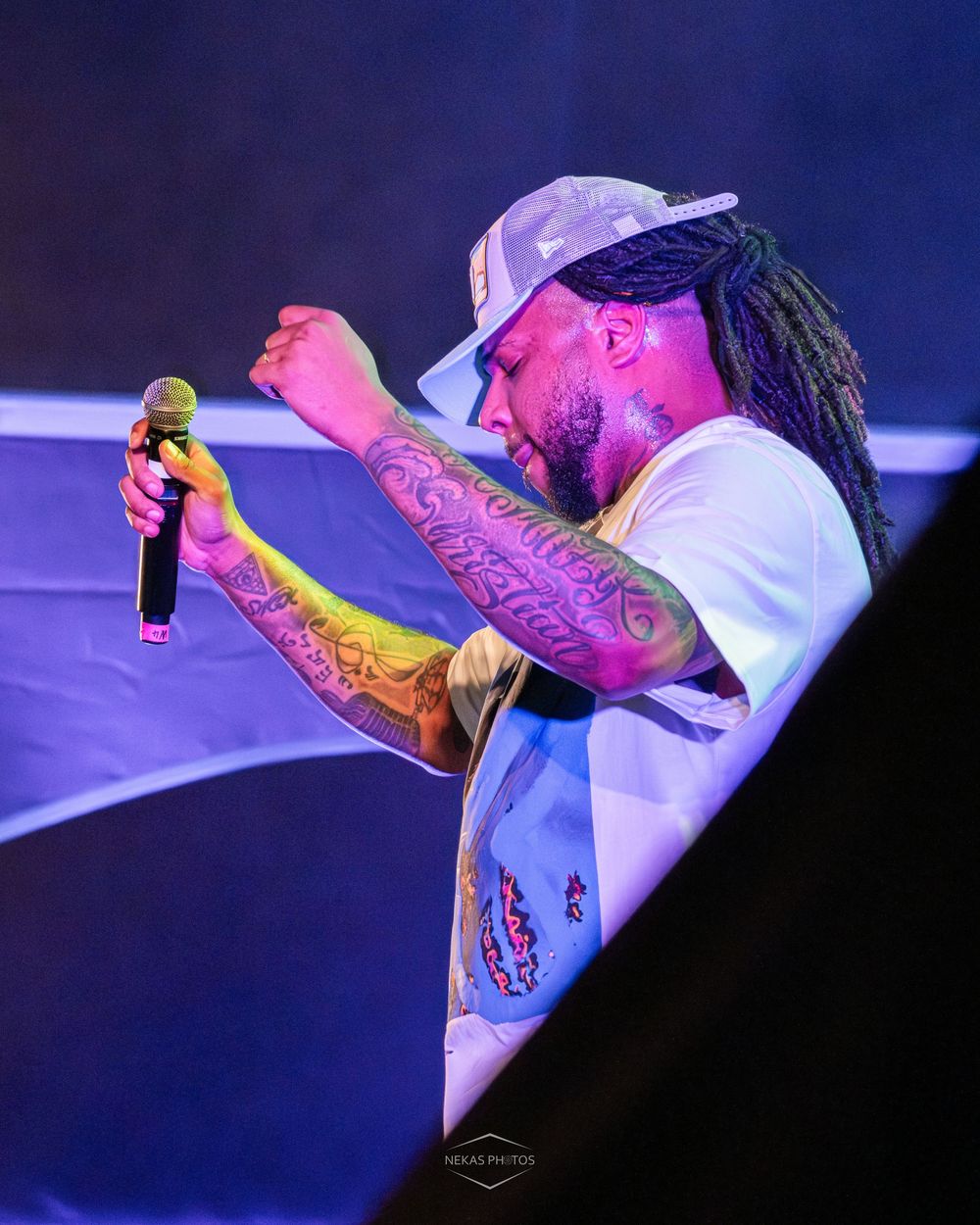 Tattooed rapper with dreadlocks and white cap performs on stage holding microphone under colorful blue and purple lights.