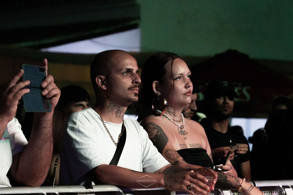 A man and woman with tattoos watch a live event, man holding a smartphone, standing in a dimly lit outdoor venue.