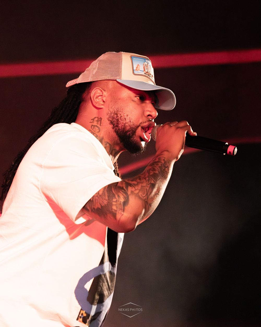Tattooed male rapper performing on stage, singing into microphone wearing white tee and gray trucker hat under red stage lights.