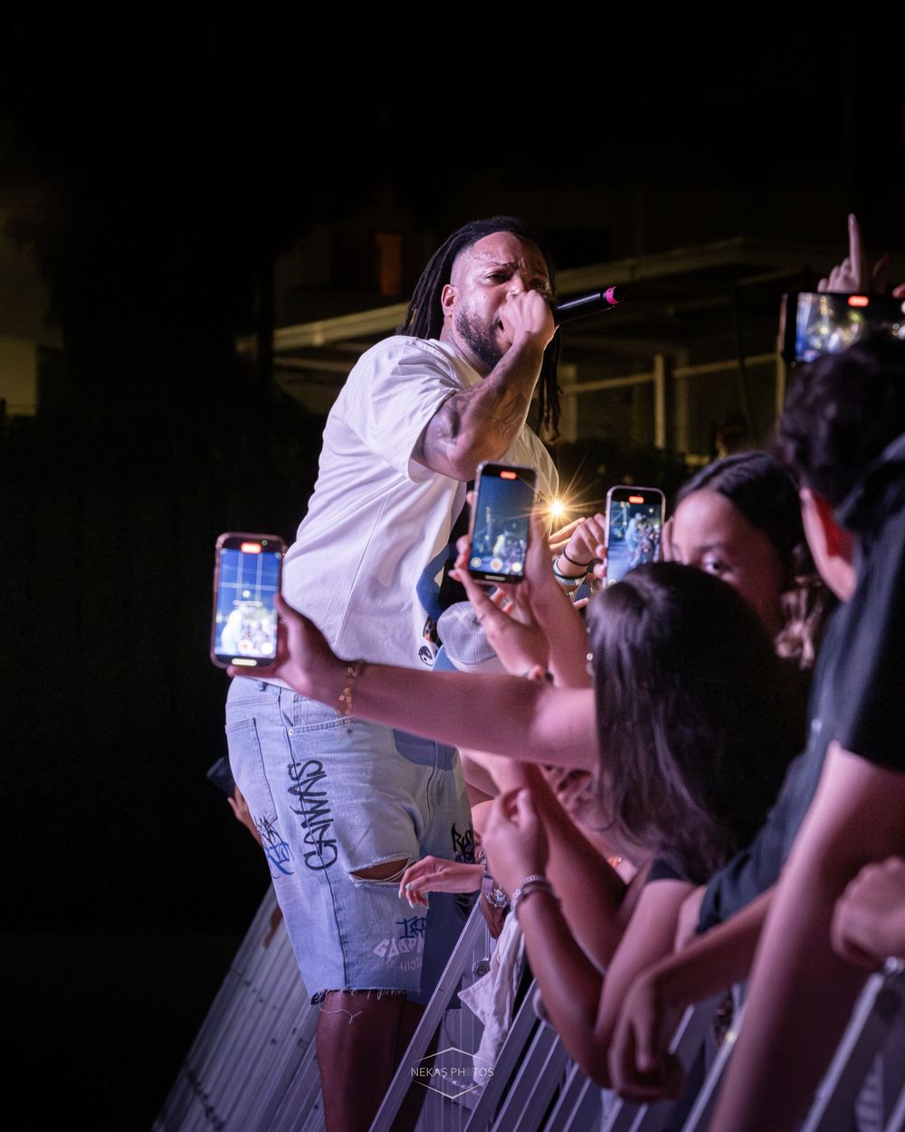 Tattooed rapper performs on stage leaning toward crowd of fans recording with smartphones at night concert.