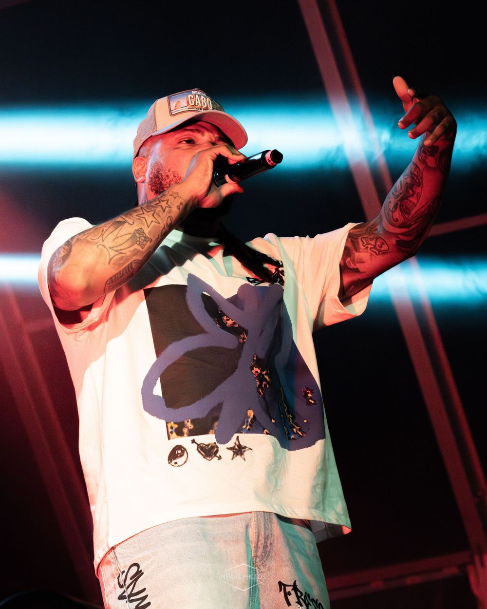 Tattooed rapper performing on stage wearing a white graphic tee and trucker hat, holding a microphone at a concert.