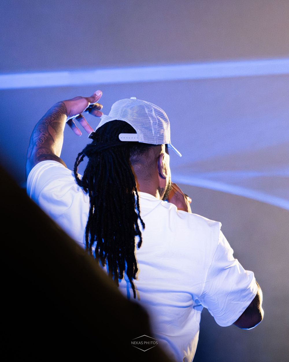 Person with dreadlocks wearing a white cap and shirt performing on stage under blue lighting.