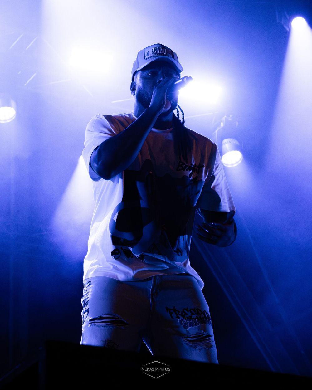 A rapper performs on stage under dramatic blue lighting, holding a microphone while wearing a white tee and ripped jeans.