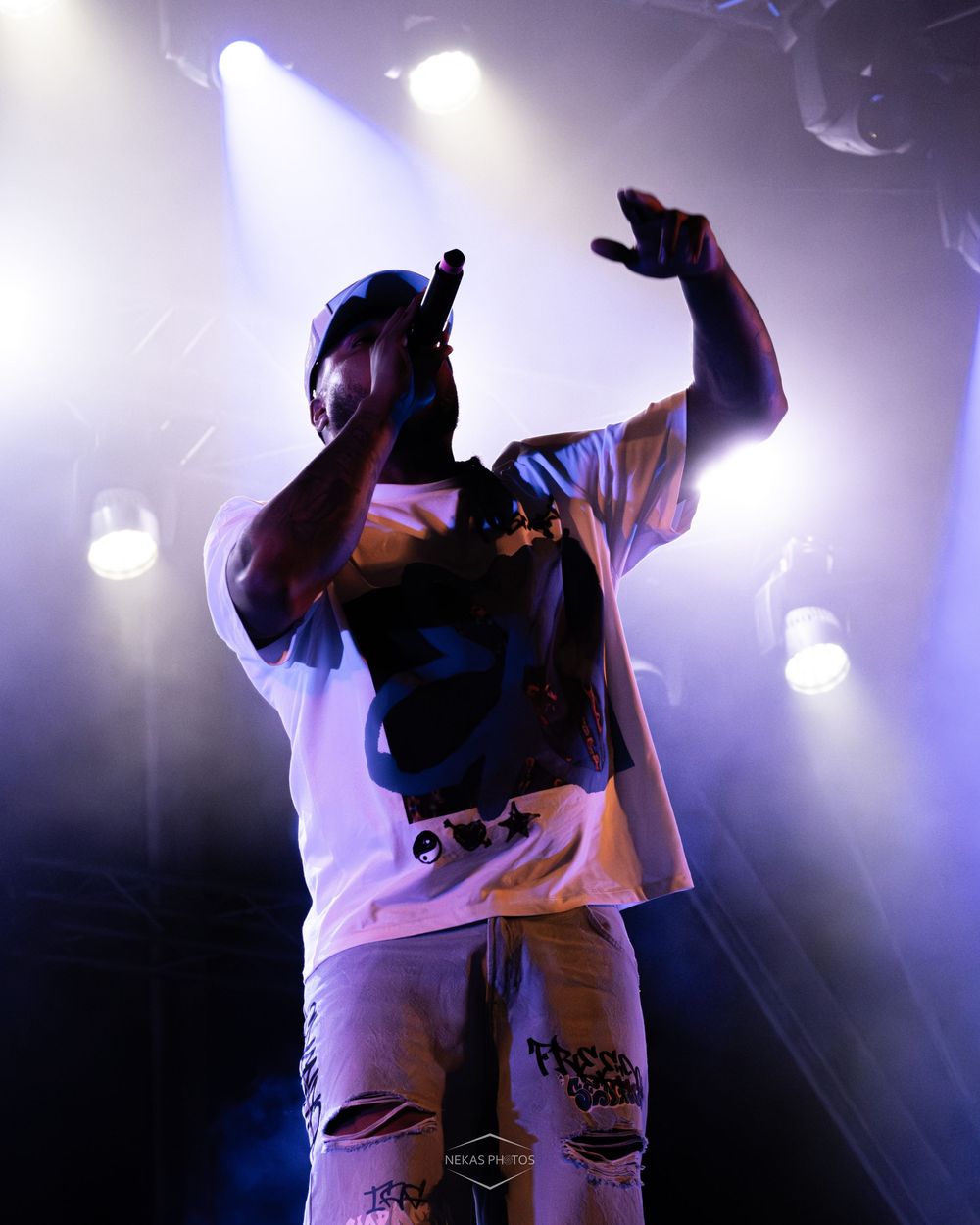A rapper performs on stage under purple lights, holding a mic and gesturing, wearing ripped jeans and a graphic tee.