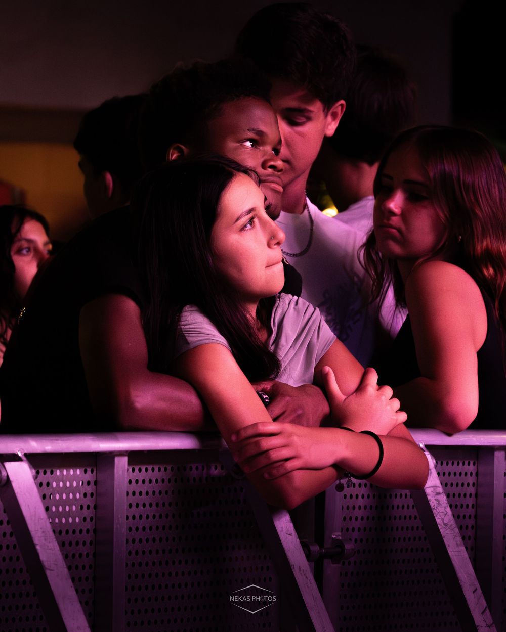 Young concert-goers lean against a barrier, bathed in purple stage lighting at a live music event.