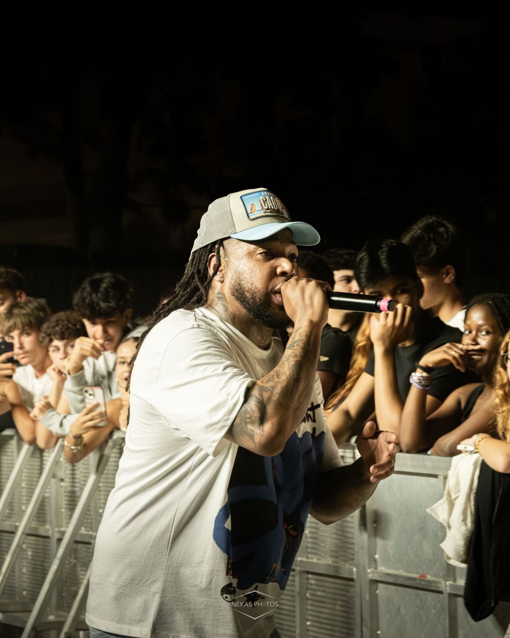 A rapper with dreadlocks and a gray cap performs into a microphone at a live concert with fans behind a barrier.