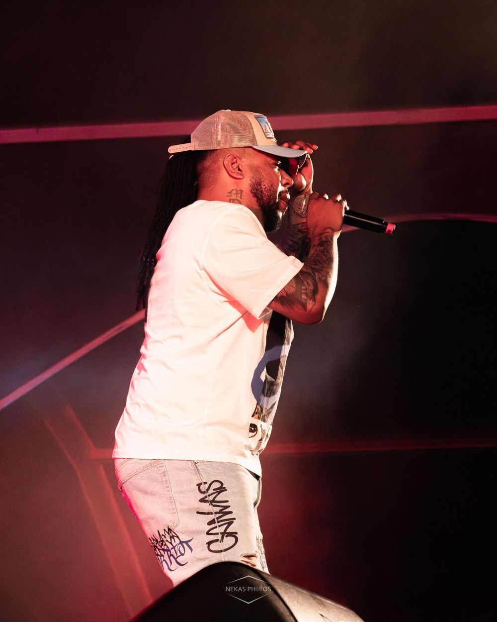 Tattooed male rapper performing on stage, holding microphone, wearing white cap and outfit under dramatic red lighting.