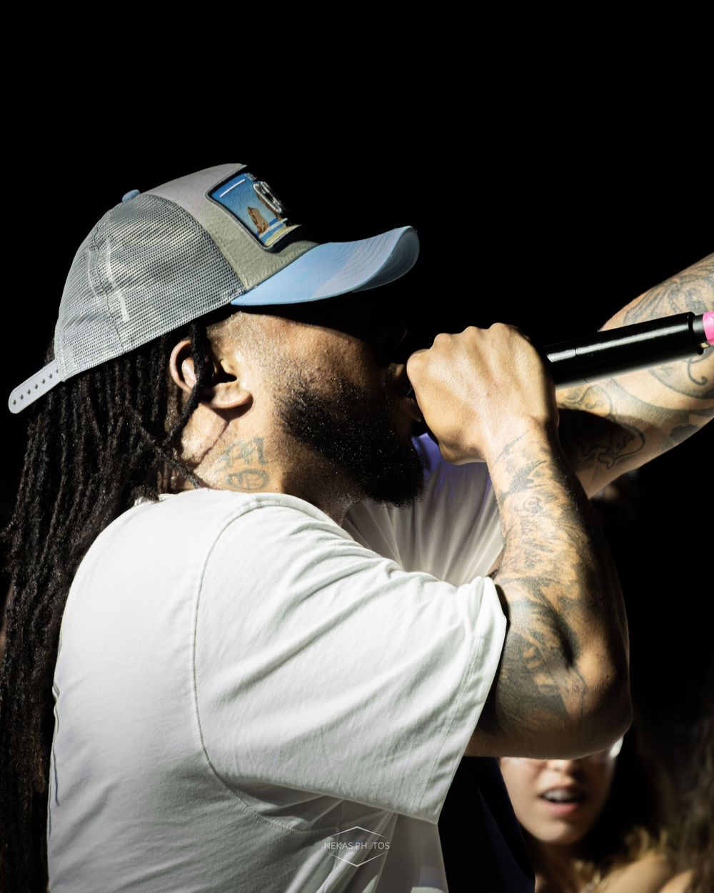 Tattooed rapper performing live on stage holding a microphone wearing a grey cap and white t-shirt.