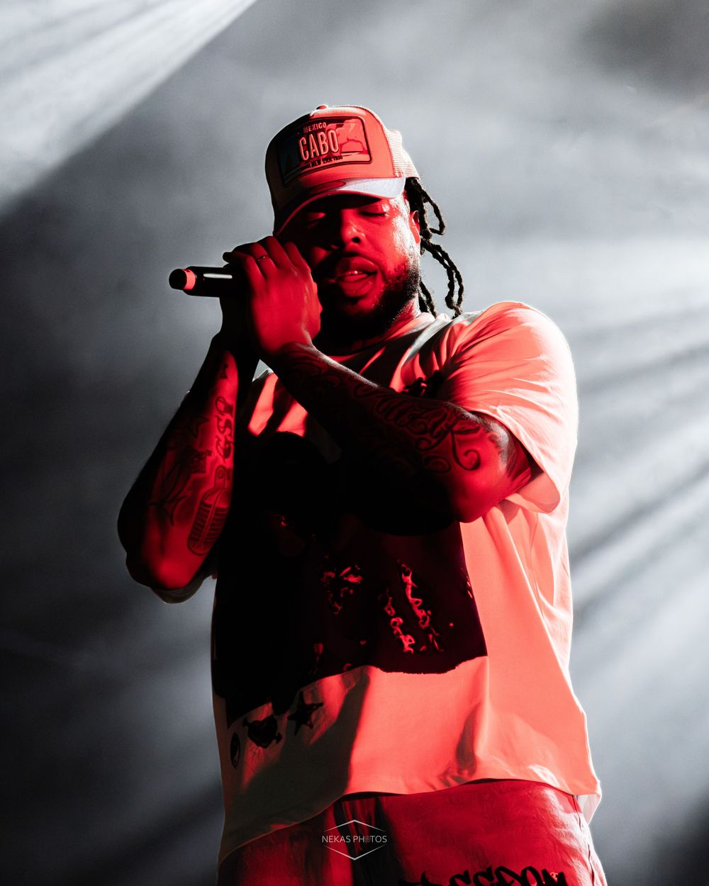 Tattooed rapper performing on stage holding microphone wearing red cap and white t-shirt under dramatic red stage lighting.