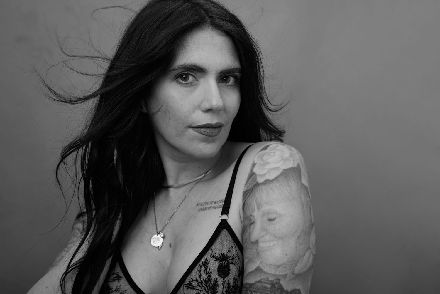 Black and white portrait of a tattooed woman with dark flowing hair wearing a floral lace top.