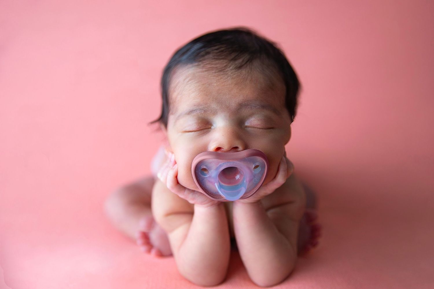 Sleeping newborn baby with pacifier posed on pink background in adorable froggy position for professional photo.