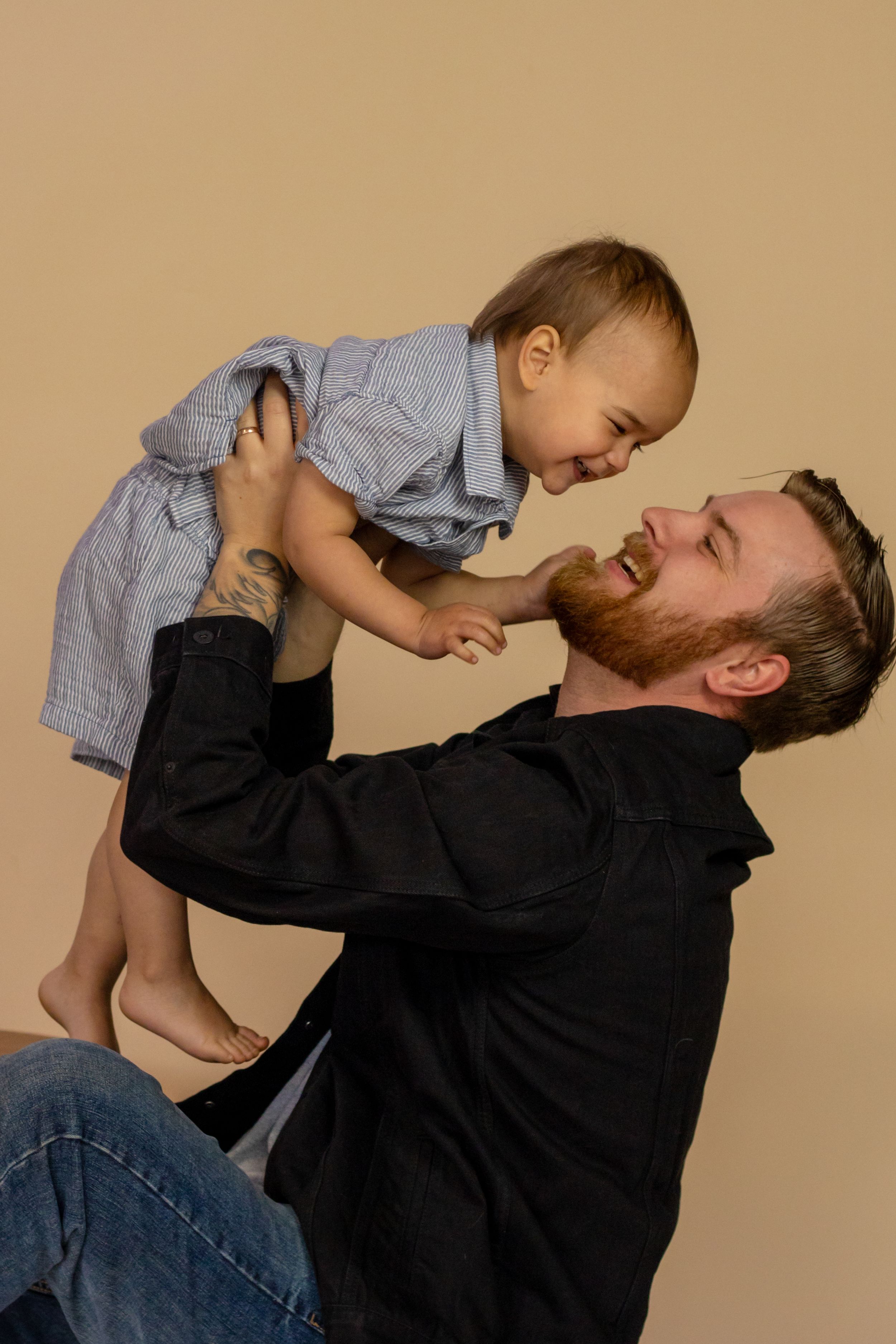 Bearded father lifting laughing toddler in striped outfit while sitting on beige studio floor in roy washington