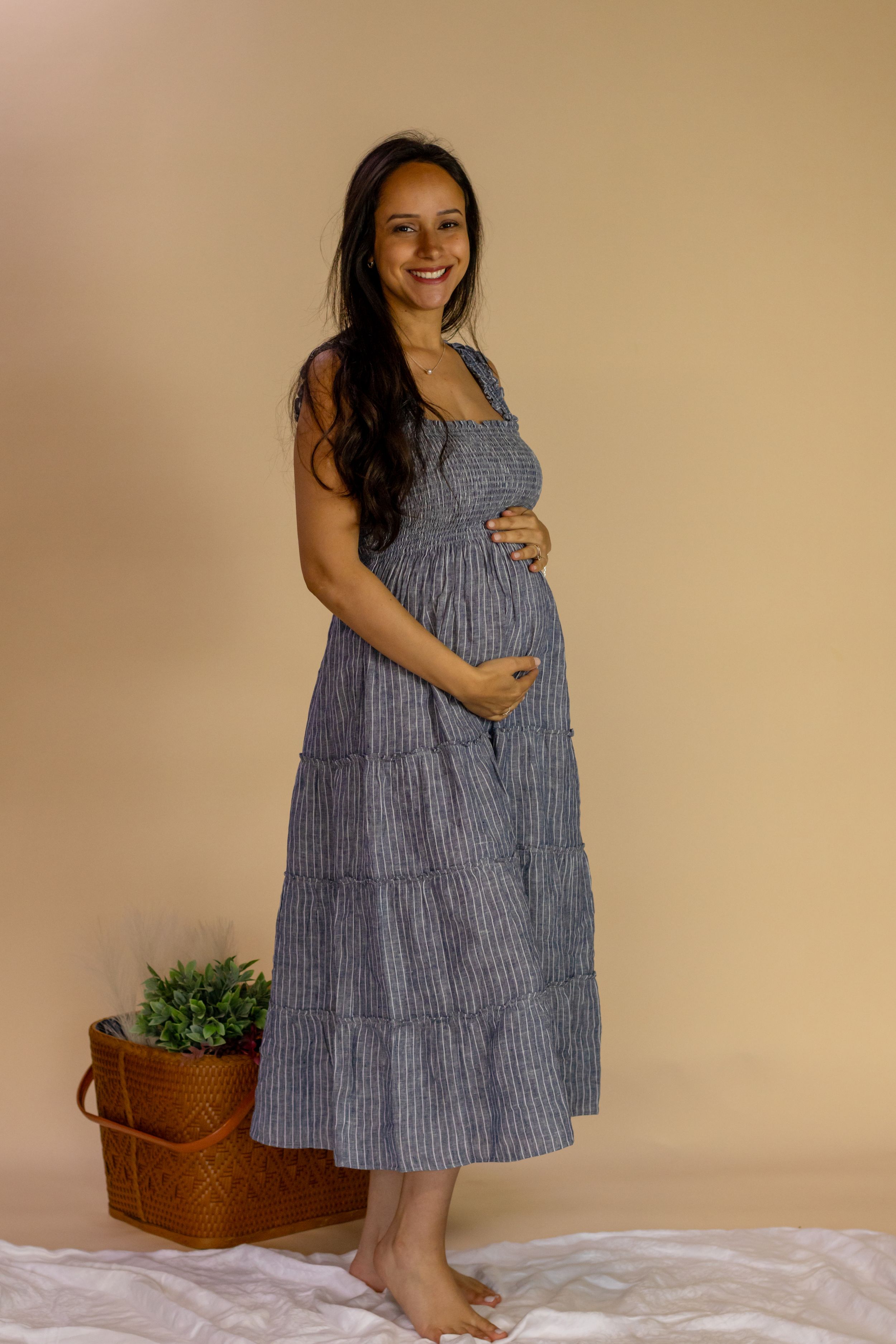 Smiling pregnant woman in blue striped sundress cradling baby bump in beige photography studio in roy washington with photographer