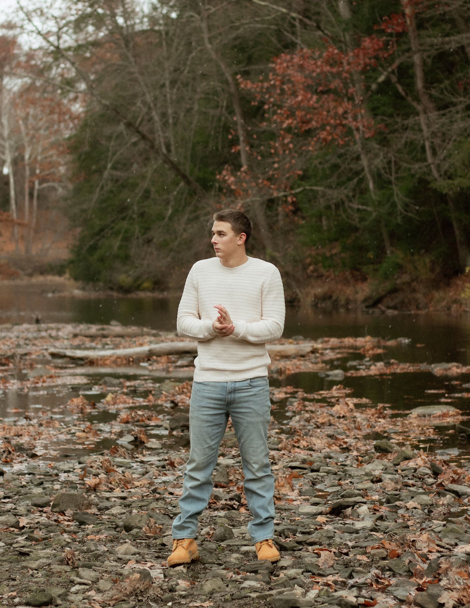 Young man in white sweater and jeans standing on rocky riverbank surrounded by autumn foliage.