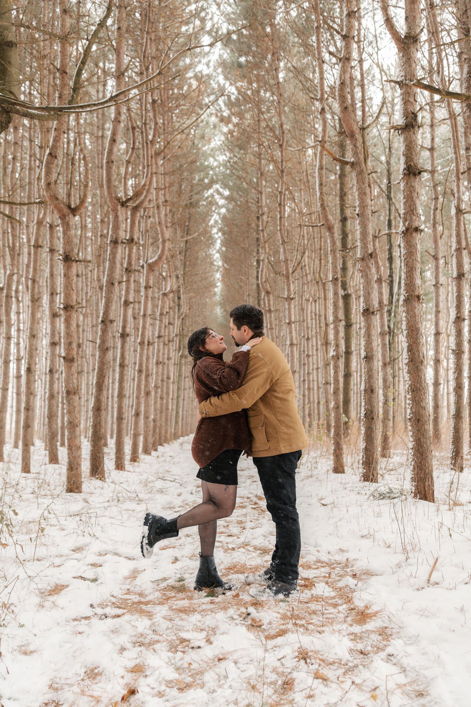 Romantic couple kissing in snowy winter forest with tall bare trees lining a snow-covered path.