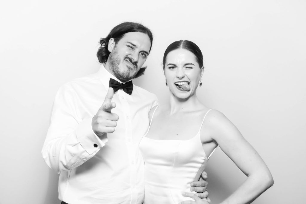 Black and white photo booth shot of a couple in formal attire, man in bow tie pointing, woman sticking out tongue.