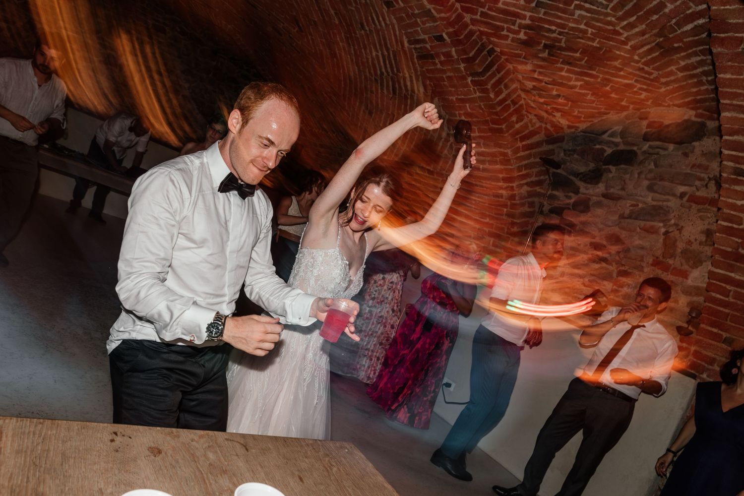 Bride and groom dancing energetically in a rustic brick venue with motion blur and colorful lights.