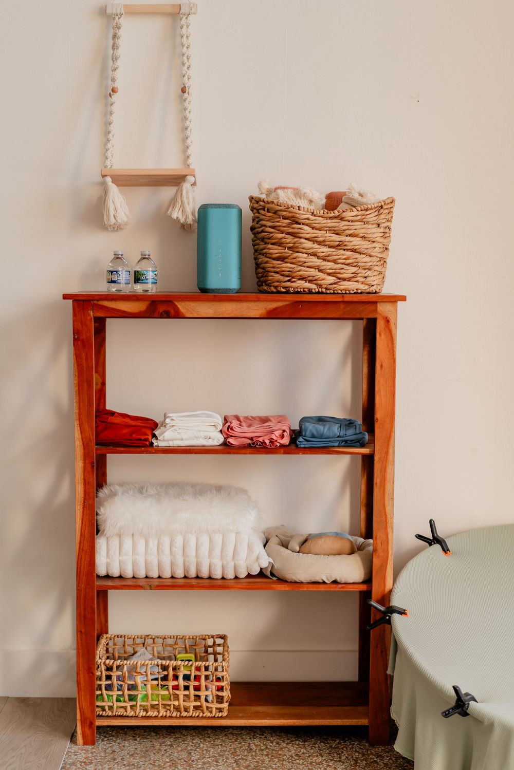 Wooden bookshelf with folded clothes, wicker baskets, and baby items against a beige wall with a decorative rope ladder.