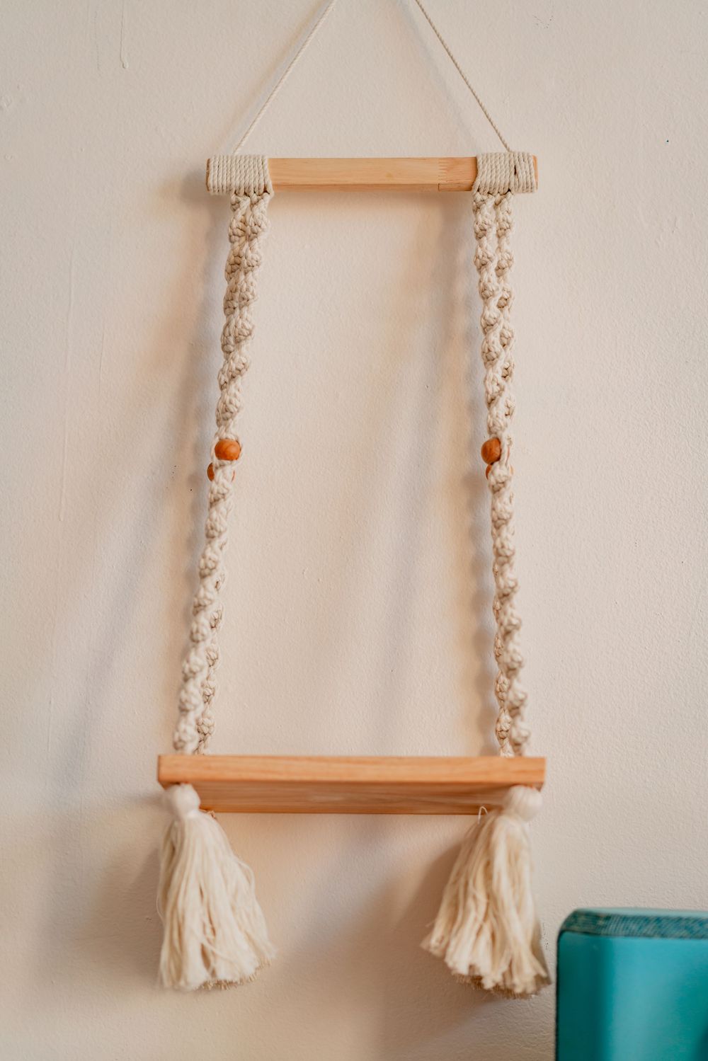 Boho macrame wall hanging shelf with wooden dowels, cream tassels, and orange beads on a white wall.