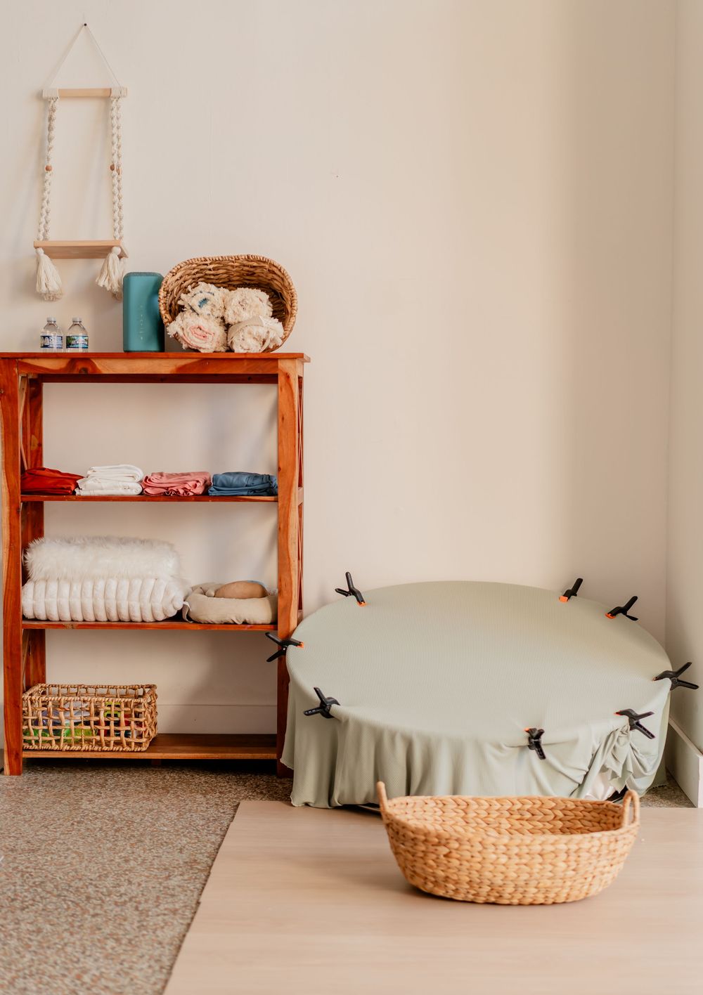 Wooden nursery shelf with folded linens, wicker baskets, and a sage green round changing pad on the floor.