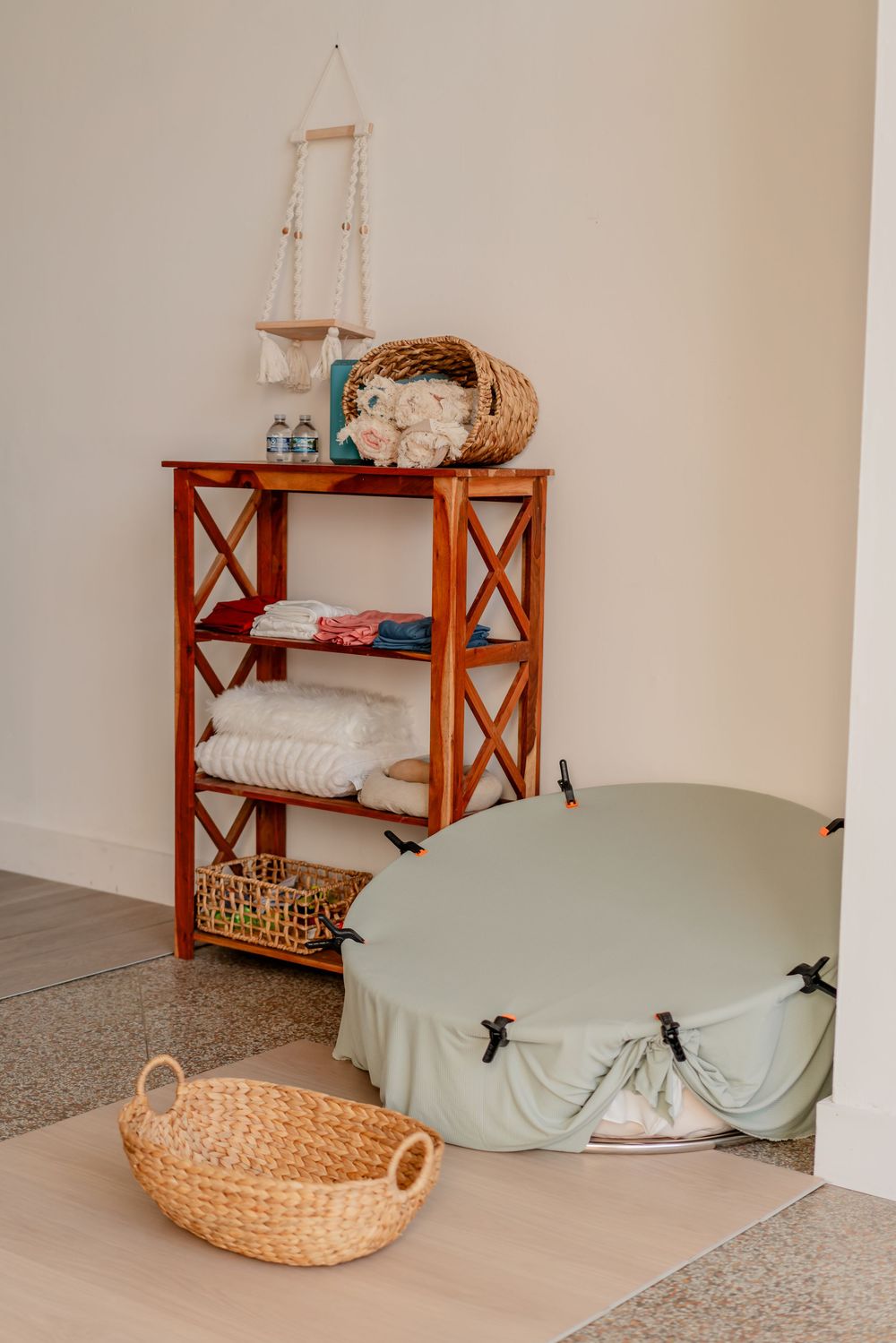 Wooden shelf with folded towels, wicker baskets, and a sage green floor cushion in a cozy, neutral-toned room.