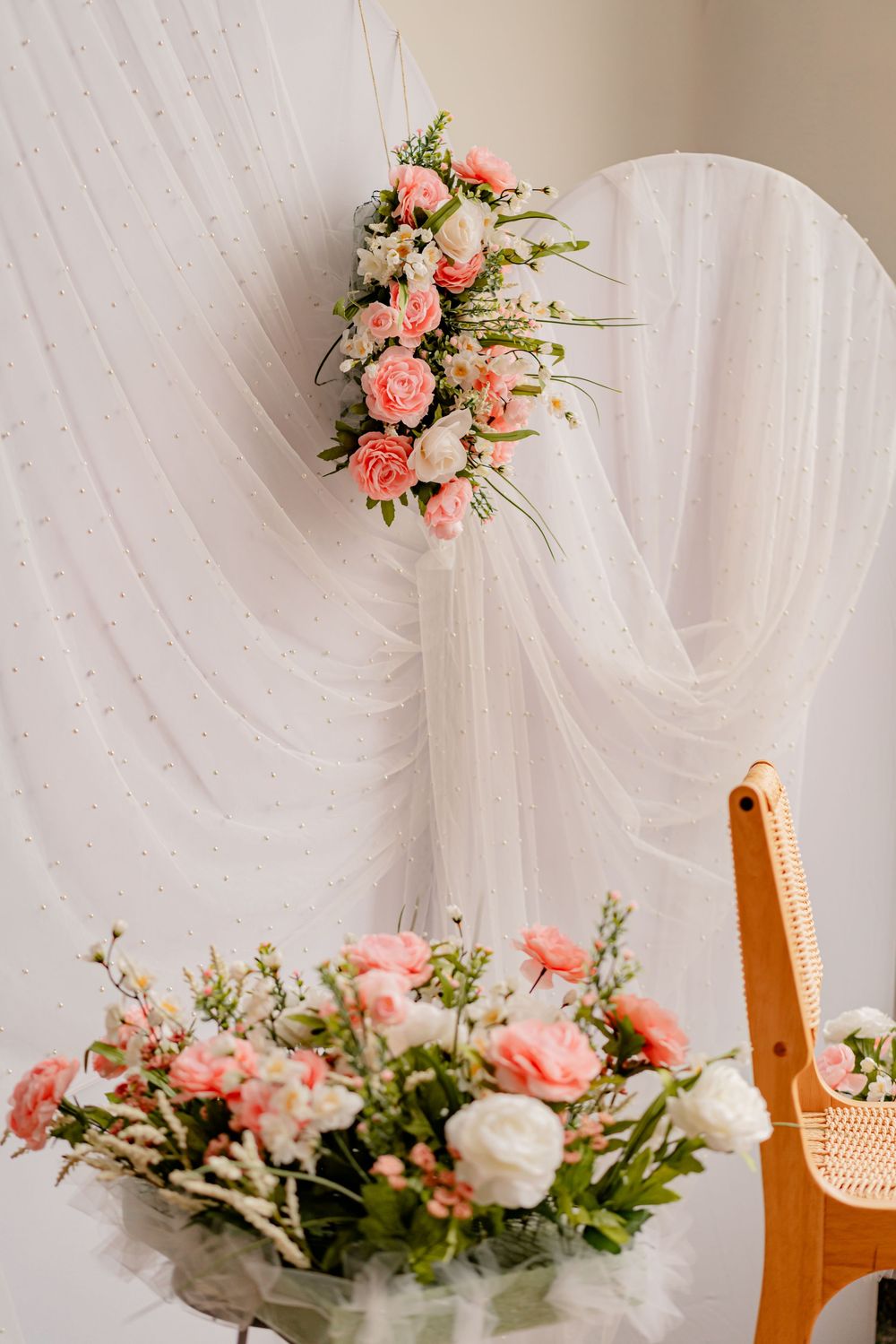 Elegant wedding decor with pink and white roses arranged on a draped white tulle backdrop with a wooden chair.