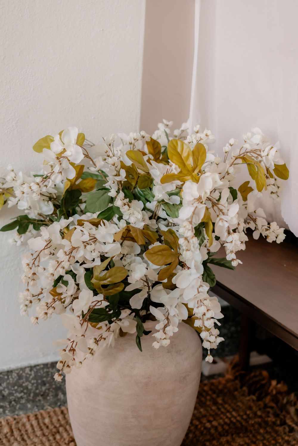 Large ceramic vase with white flowering branches and green leaves displayed on a woven rug beside a wooden table.
