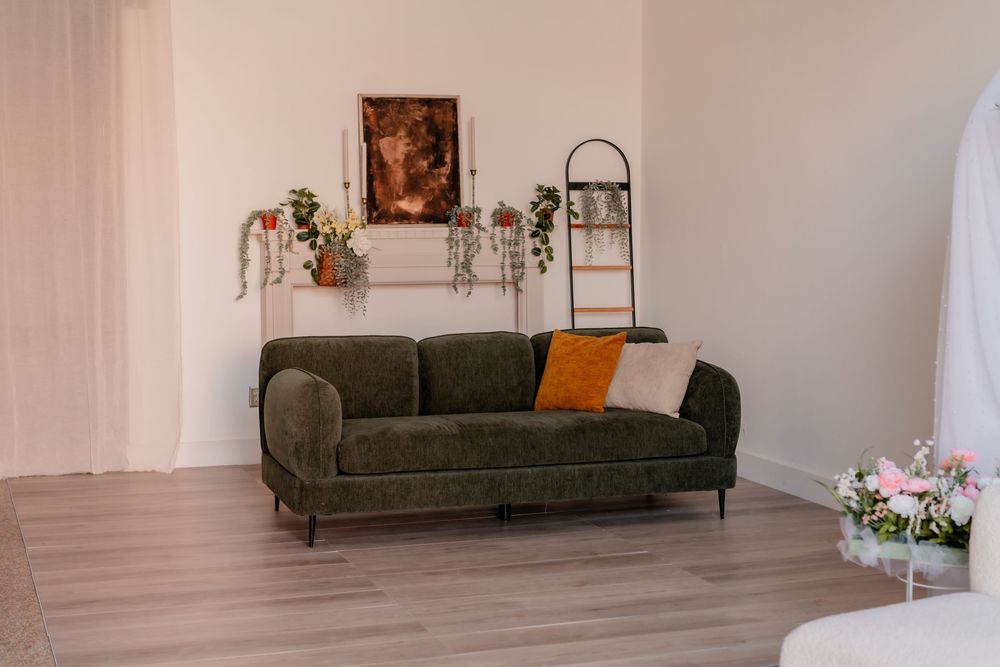 Green velvet sofa with orange pillow in a bright living room, decorated with plants and artwork on a shelf.