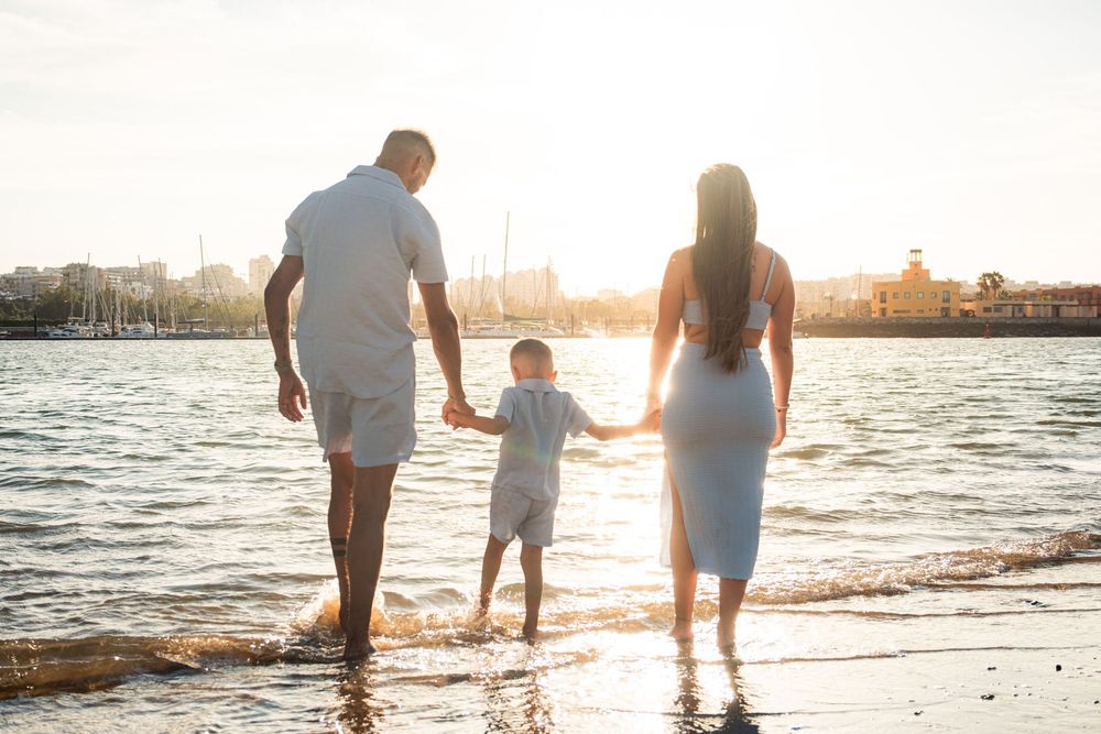 Family of three holding hands walking along a sunny beach at golden hour with calm water and marina in background.