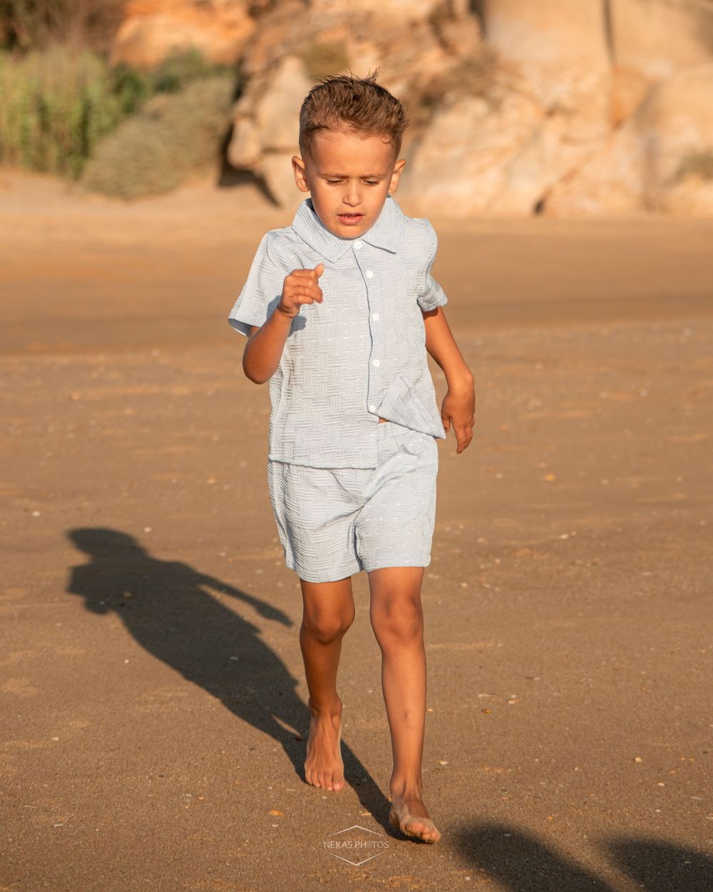 Young boy in light blue linen shorts set walking barefoot on sandy beach with rocky cliffs in background.
